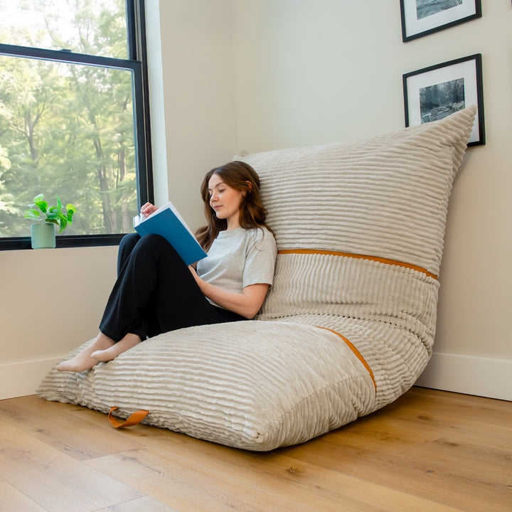 A woman sits on a large ribbed wedge floor cushion in a bright room, reading a blue book. The cushion is upholstered in a light beige textured fabric with subtle orange-brown accent stripes and is positioned against a wall near a window. 