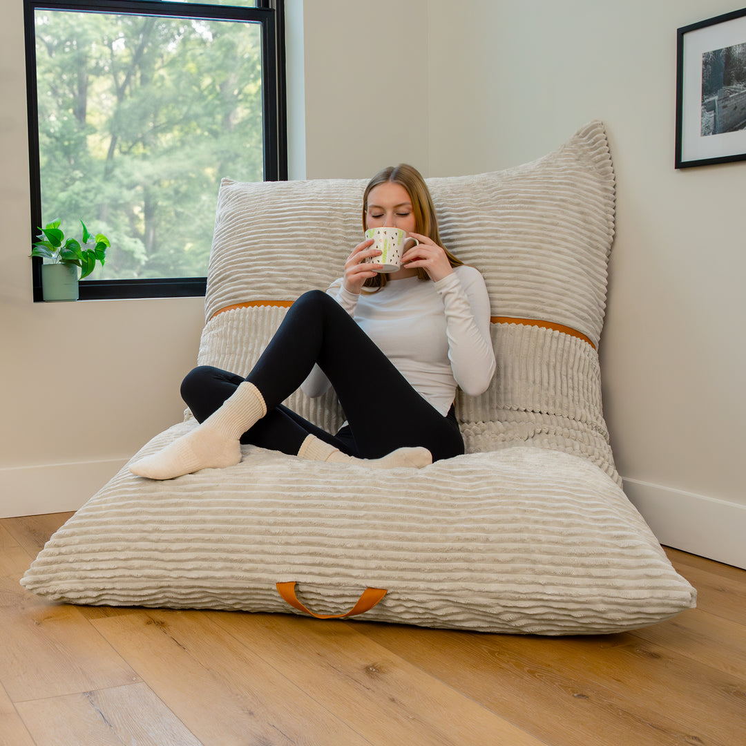 A woman sits cross-legged on a large ribbed floor cushion with a matching backrest in a bright room with a window, wood floor, and framed wall art. 