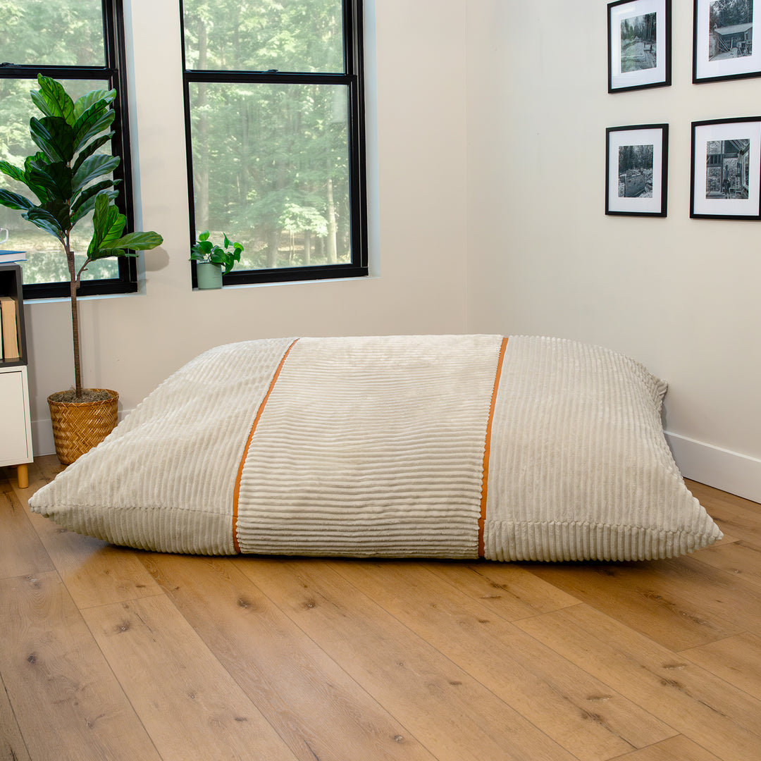 A large rectangular floor cushion or mattress topper in a cream corduroy fabric with two vertical orange stripes, resting on a light wood floor in a modern room. 