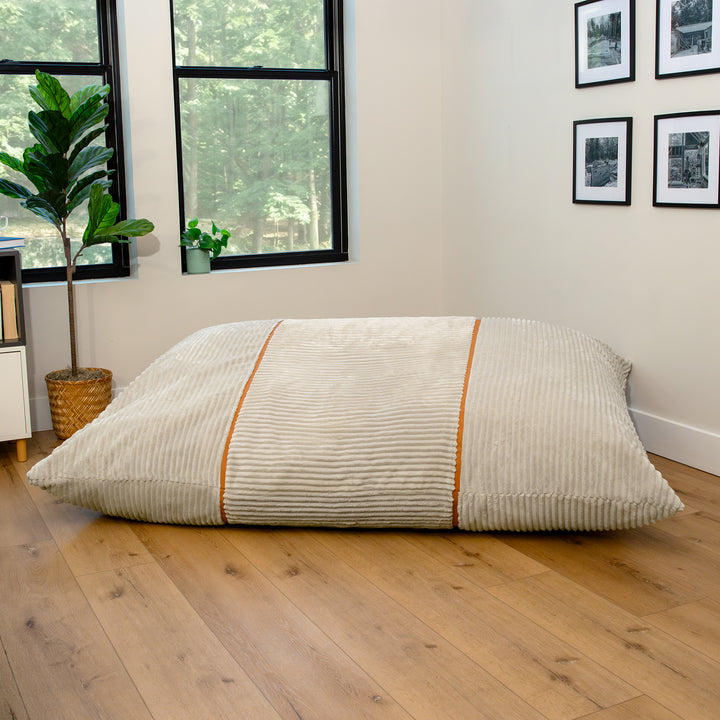 A large rectangular floor cushion or mattress topper in a cream corduroy fabric with two vertical orange stripes, resting on a light wood floor in a modern room. 