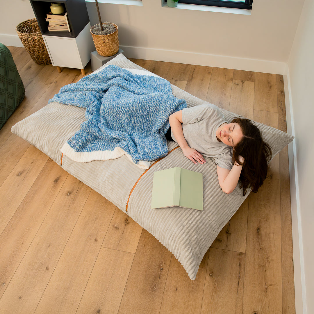 A person lies on a large striped floor cushion in a bedroom, covered with a blue blanket and reading a light green book. The room has light wood flooring and neutral walls. 