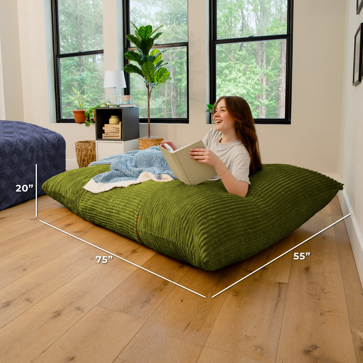 A woman relaxes on a large olive green ribbed floor cushion in a bright bedroom, reading a book. White dimension lines overlay the cushion showing measurements of 75 inches by 55 inches by 20 inches. 