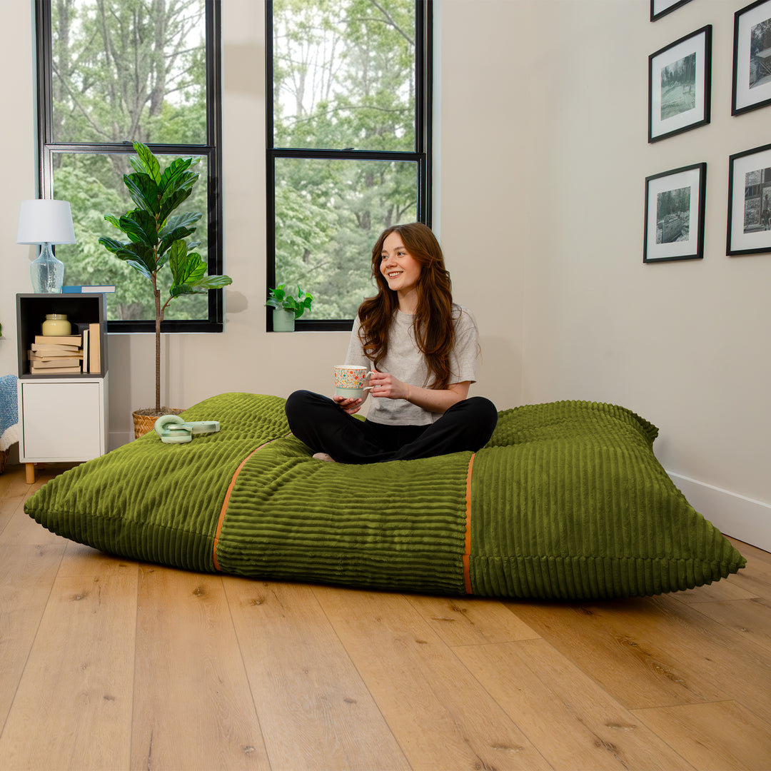 A woman sits cross-legged on a large olive green ribbed floor cushion in a bright room with wood floors, large windows, and framed wall art. 