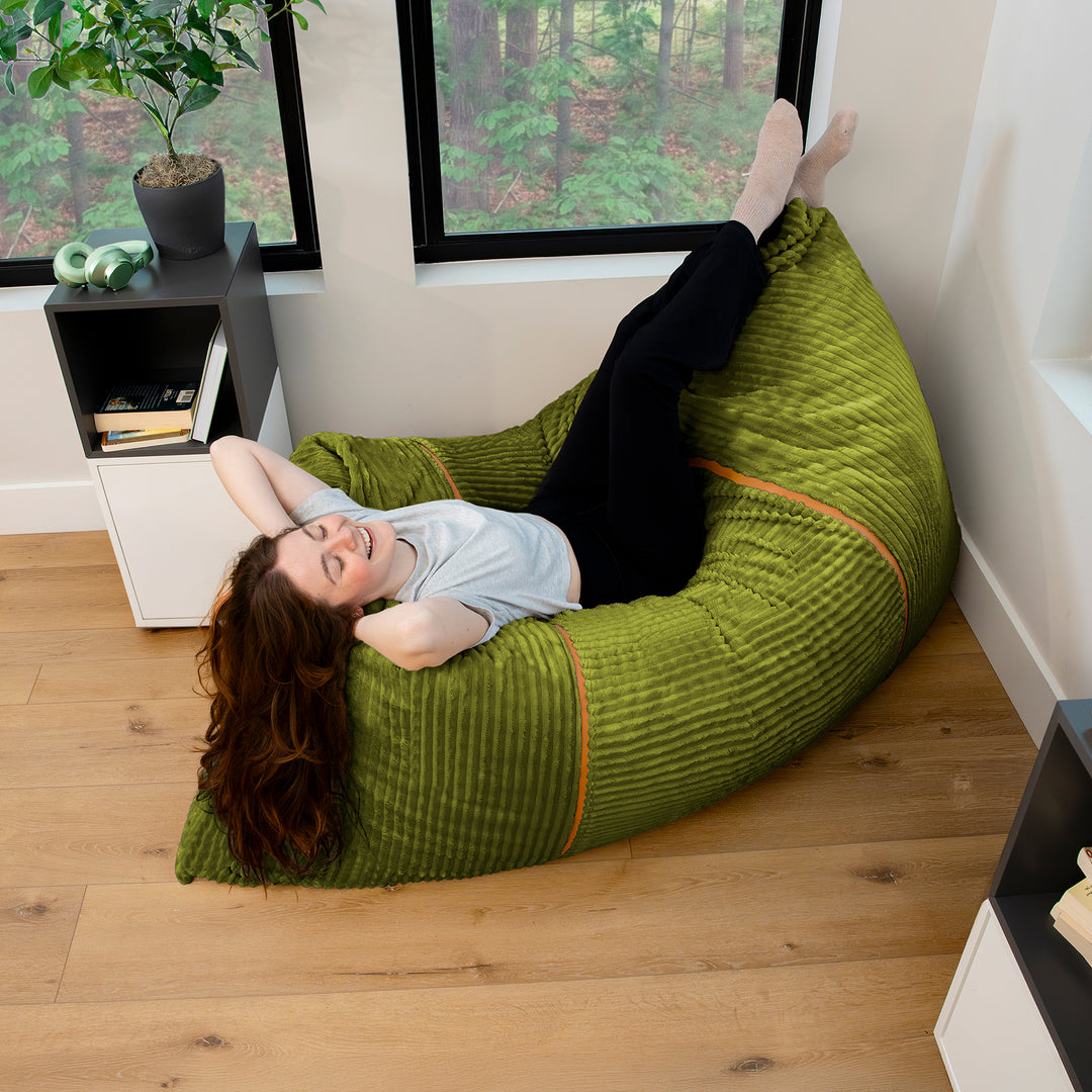 A woman lies on a large olive green ribbed bean bag chair in a bright room with wood floors and windows. 