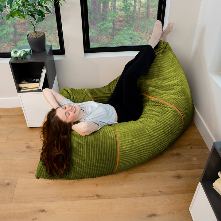 A woman lies on a large olive green ribbed bean bag chair in a bright room with wood floors and windows. 