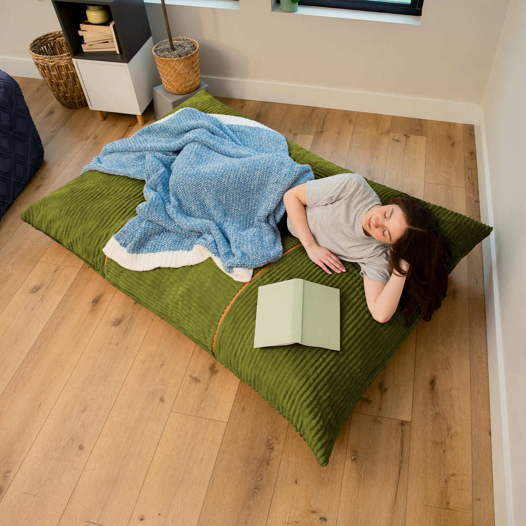 A person lies on a large green ribbed floor cushion or mattress pad on a light wood floor, with a blue blanket draped over them and an open book nearby in a bedroom setting. 