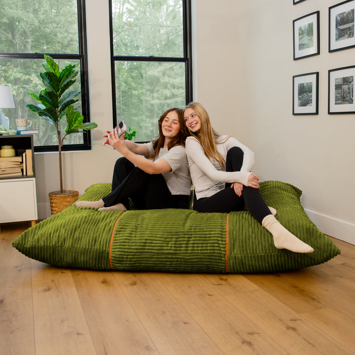 Two women sitting on a large olive green ribbed floor cushion in a bright, modern room with wood floors and large windows. 