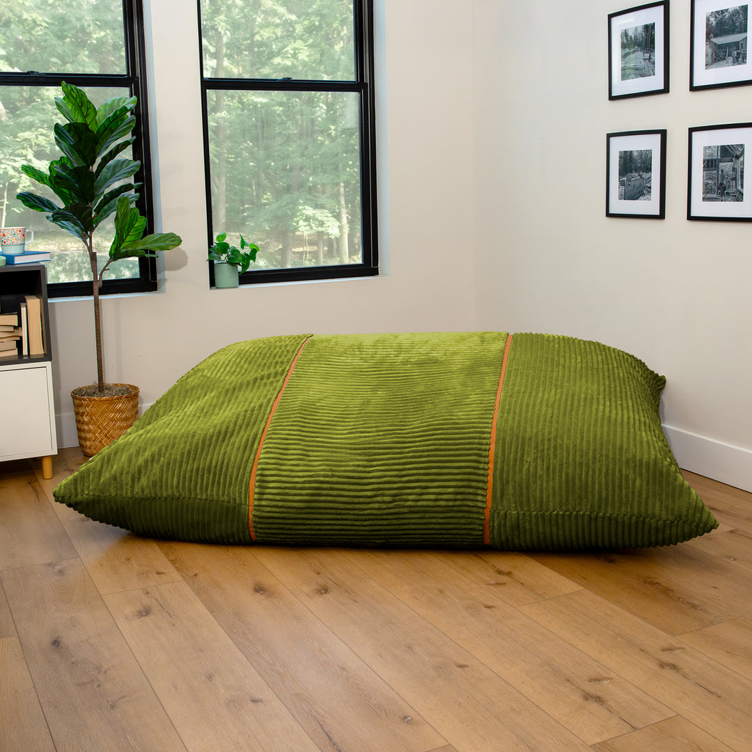 A large olive green corduroy floor cushion or bean bag with orange piping details, displayed on a light wood floor in a modern room setting. 