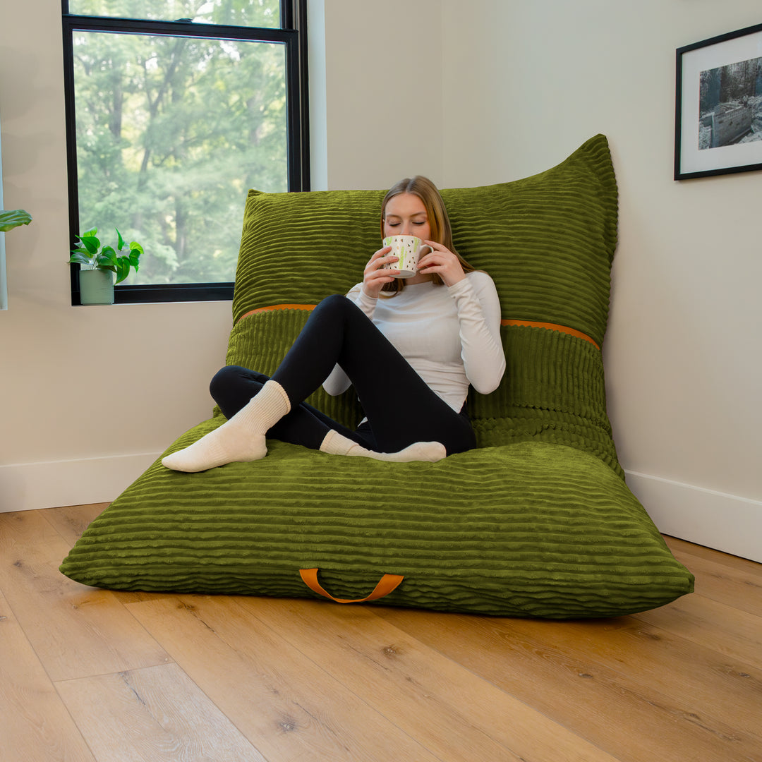 A woman sits cross-legged on a large olive green ribbed floor cushion with a matching back pillow in a bright room with a window and wood floor. 