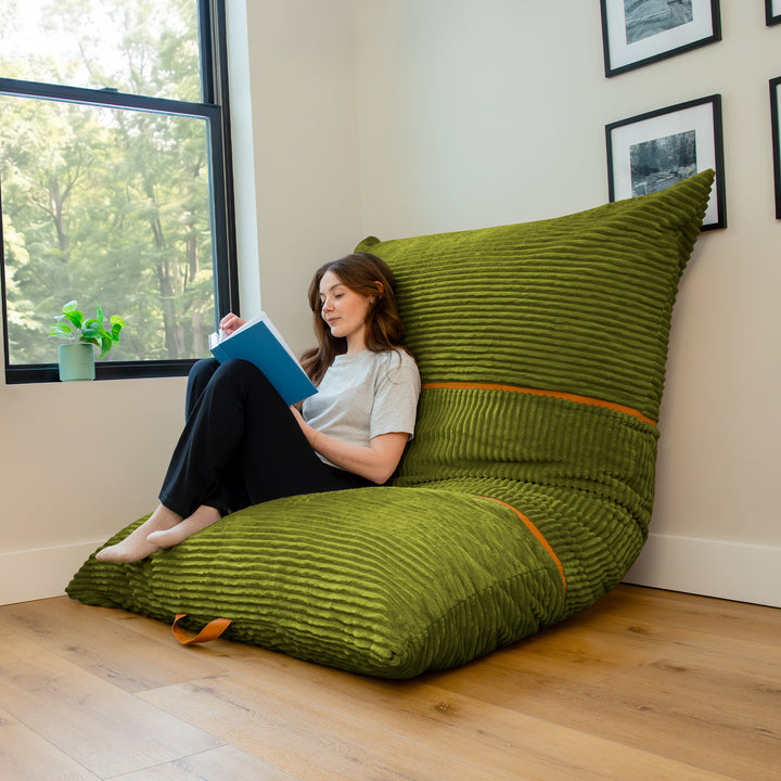 A woman sits on a large olive green ribbed lounge chair in a bright room with a window, reading a blue book. 