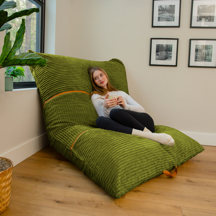 A woman reclines on a large olive green corduroy floor chair in a bright room with wood floors, framed wall art, and a potted plant. 