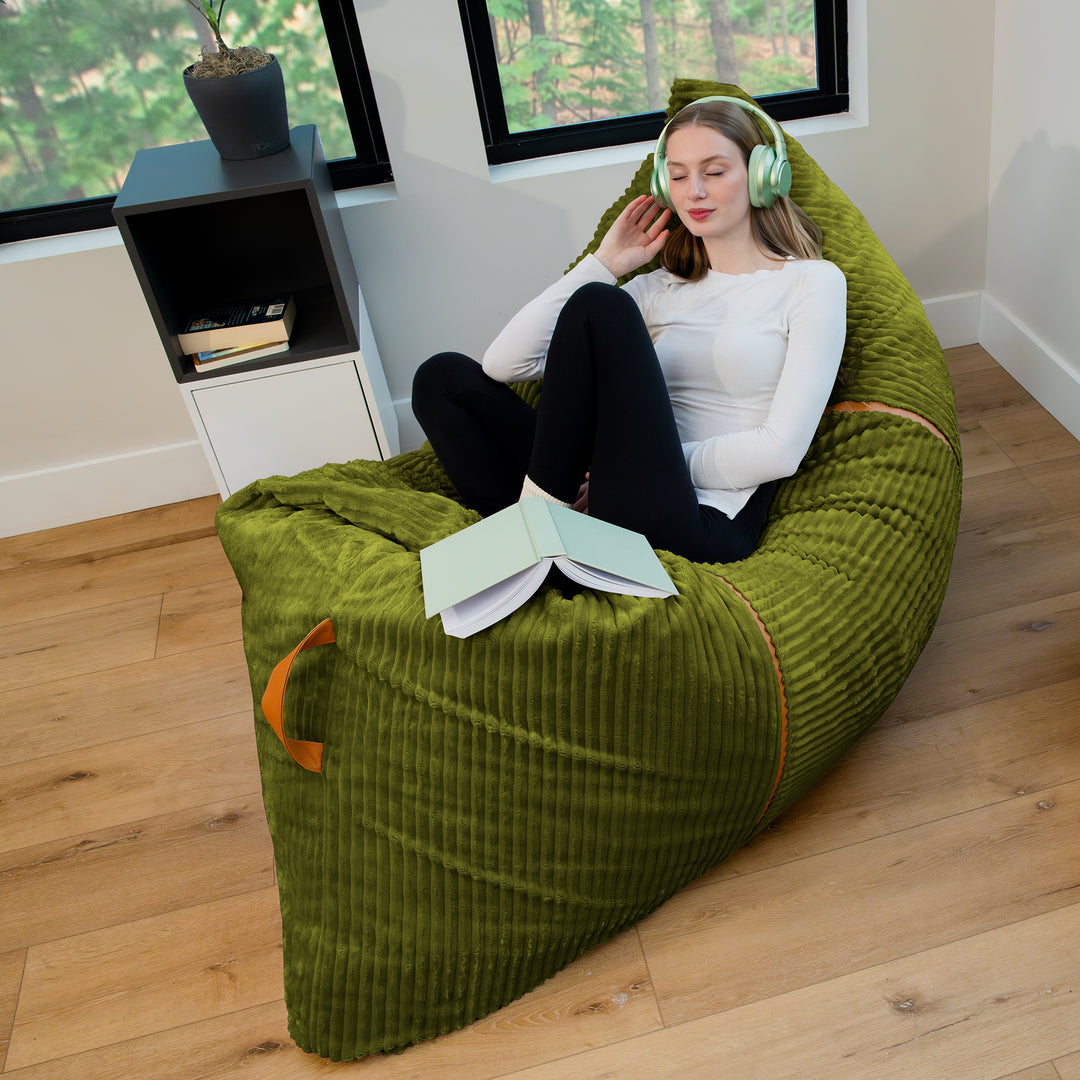 A woman sits cross-legged on a large olive green corduroy bean bag chair in a bright room with wood floors, white walls, and black-framed windows. She is wearing light green headphones and has an open book on her lap. 