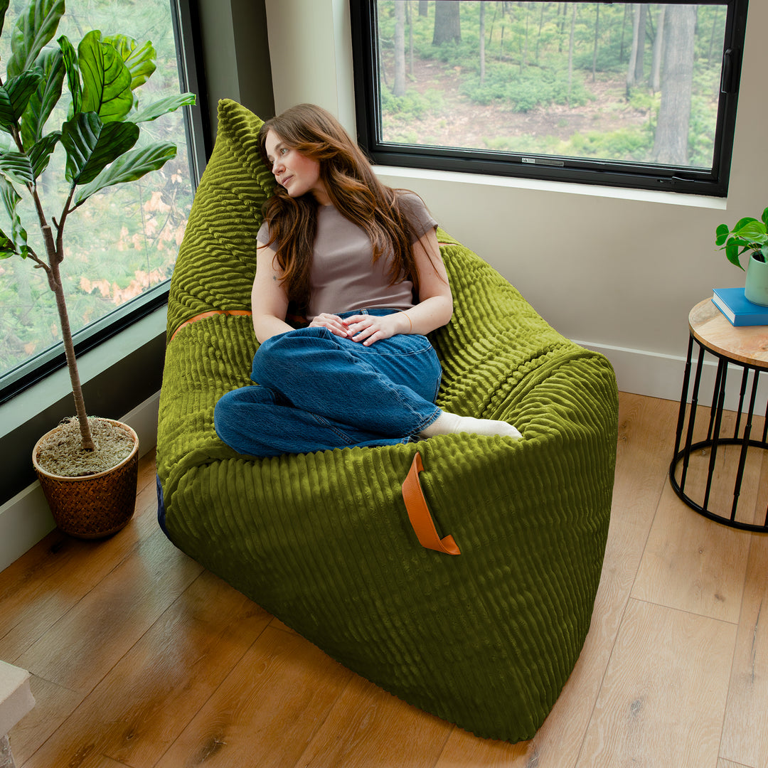 A woman sits cross-legged on a large olive green ribbed bean bag chair in a bright room with wood floors, large windows, and indoor plants. 