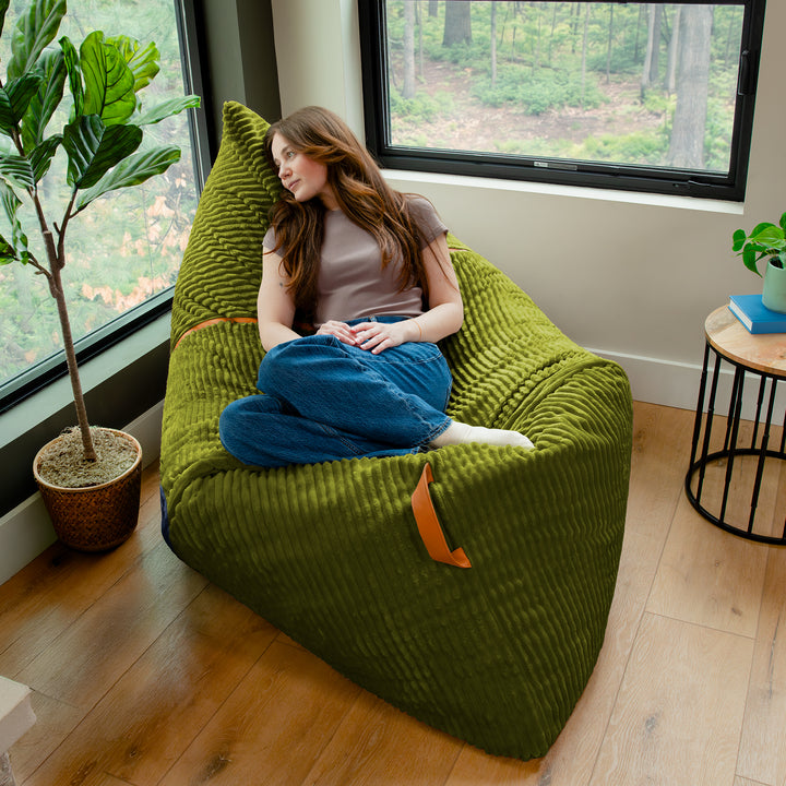A woman sits cross-legged on a large olive green ribbed bean bag chair in a bright room with wood floors, large windows, and indoor plants. 