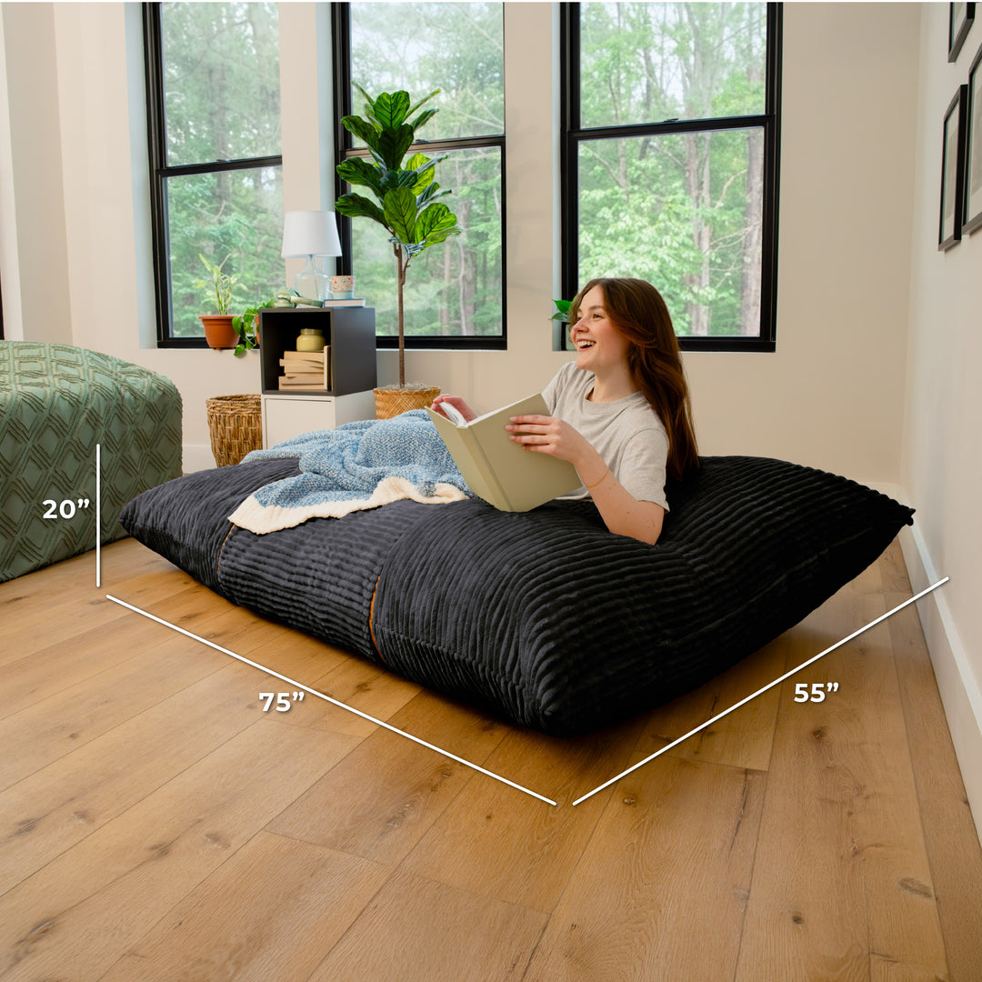A woman sits on a large black floor cushion in a bright room with wood floors and large windows. White dimension lines and text show the cushion measurements as 75 inches by 55 inches by 20 inches. 