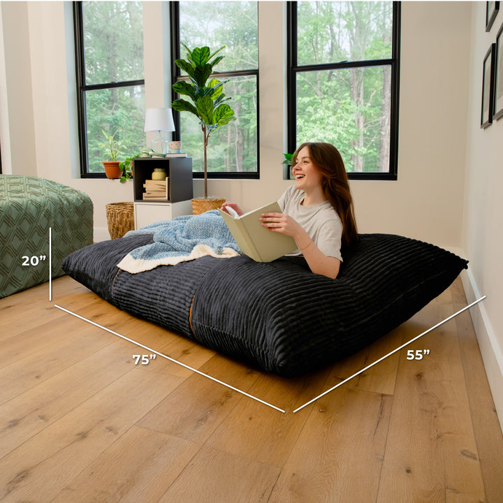 A woman sits on a large black floor cushion in a bright room with wood floors and large windows. White dimension lines and text show the cushion measurements as 75 inches by 55 inches by 20 inches. 