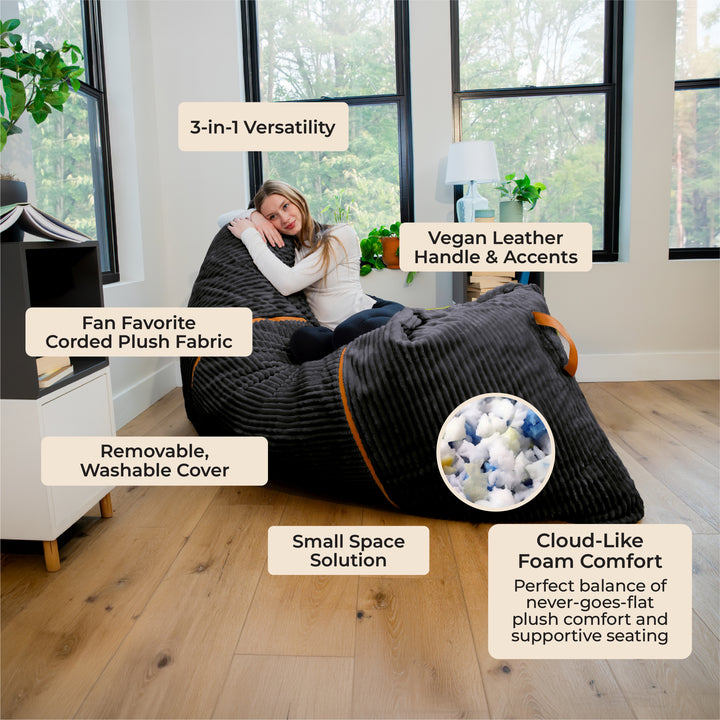 A woman reclines on a large black corduroy bean bag chair in a bright room with wood floors and large windows. Multiple text callouts describe product features, including &