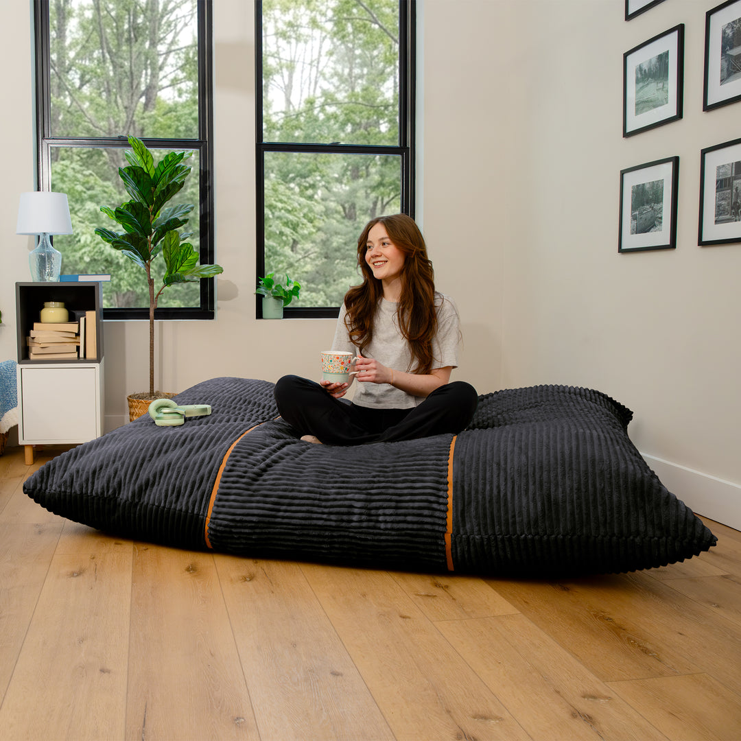 A woman sits cross-legged on a large black ribbed floor cushion in a bright room with wood floors, large windows, and framed wall art. 
