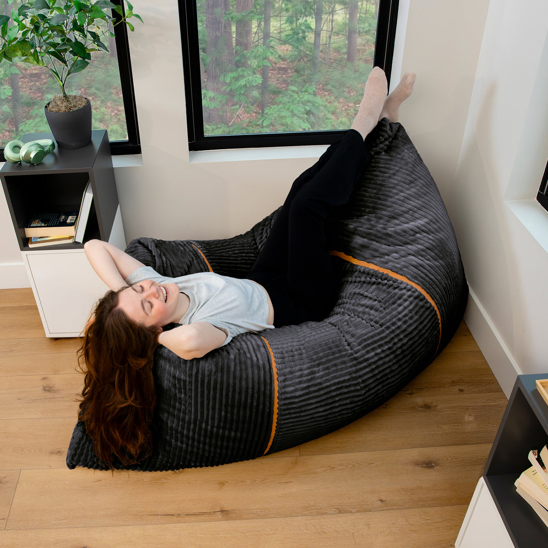 A woman relaxes in a large, curved black bean bag chair with subtle orange piping, placed on a light wood floor in a modern room with a window showing a forest view. 