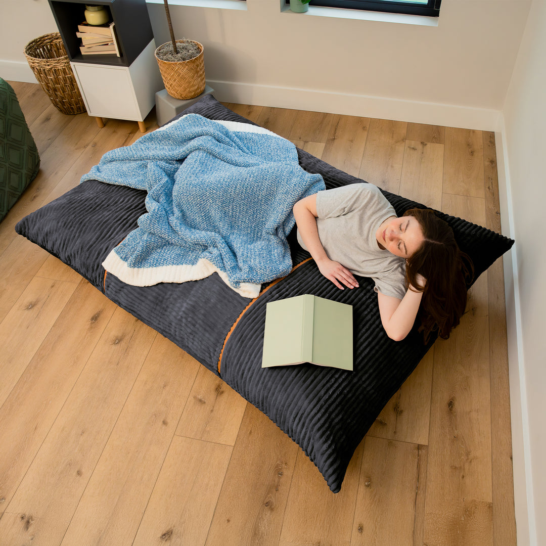 A person lies on a large dark charcoal floor cushion in a bedroom, covered with a blue textured blanket and reading a light green book. The room has light wood floors and neutral walls. 