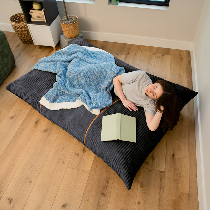 A person lies on a large dark charcoal floor cushion in a bedroom, covered with a blue textured blanket and reading a light green book. The room has light wood floors and neutral walls. 