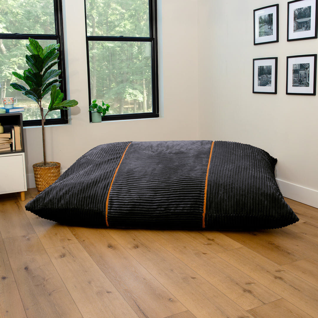 A large black ribbed floor cushion or bean bag with orange piping details, displayed on a light wood floor in a modern room setting. 
