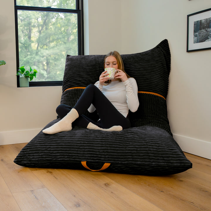 A woman sits cross-legged on a large black ribbed floor cushion with a matching back pillow in a bright room with wood flooring and a window. 