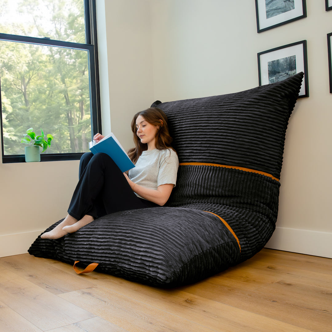 A woman sits on a large black ribbed lounge chair in a bright room with wood floors, a window, and framed wall art. The chair has a tall backrest and small orange accent bands. 