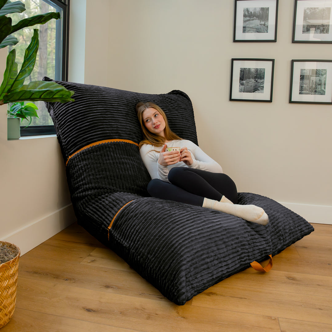 A woman sits on a large black ribbed lounge chair in a bright room with wood floors, a window, and framed wall art. 