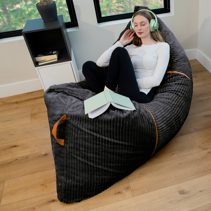 A woman sits reclined in a large dark gray ribbed bean bag chair with orange accent piping, placed on a light wood floor in a bright room. She is wearing mint green headphones and has an open book in front of her. 