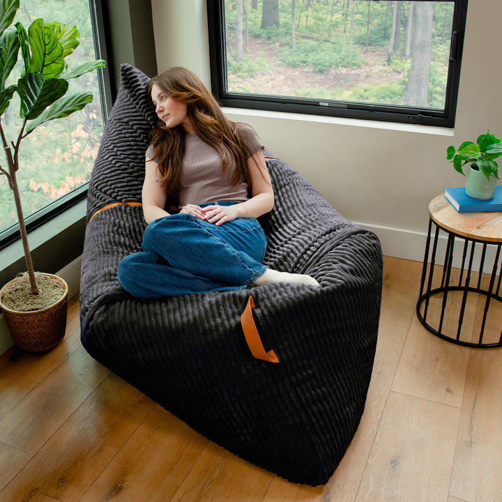 A woman sits cross-legged on a large black ribbed bean bag chair in a bright room with wood floors, large windows, and potted plants. 