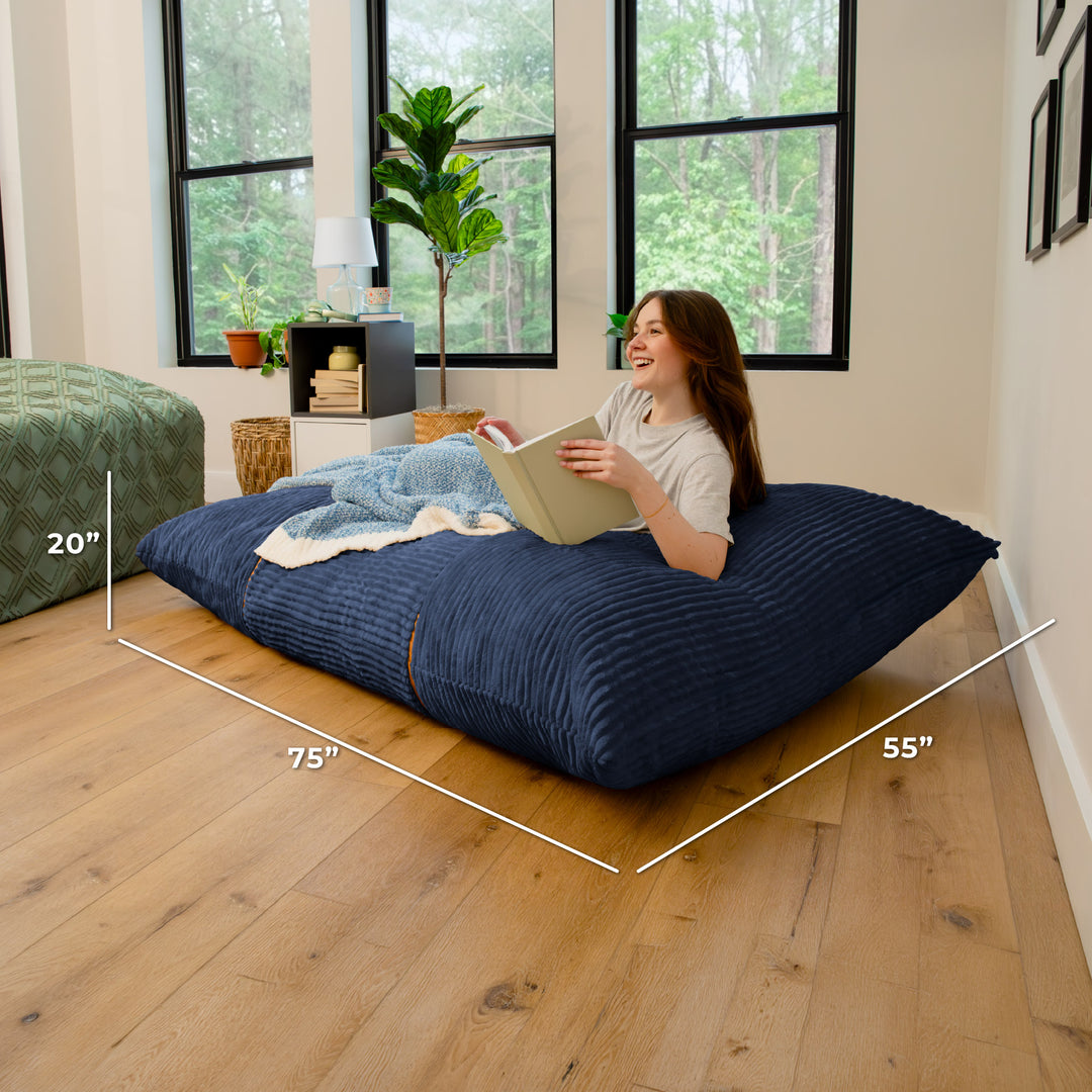 A woman reclines on a large navy blue ribbed floor cushion in a bright room with wood floors and large windows. White dimension lines overlay the cushion showing measurements of 75 inches by 55 inches, with a 20-inch height note. 