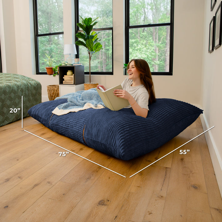 A woman reclines on a large navy blue ribbed floor cushion in a bright room with wood floors and large windows. White dimension lines overlay the cushion showing measurements of 75 inches by 55 inches, with a 20-inch height note. 