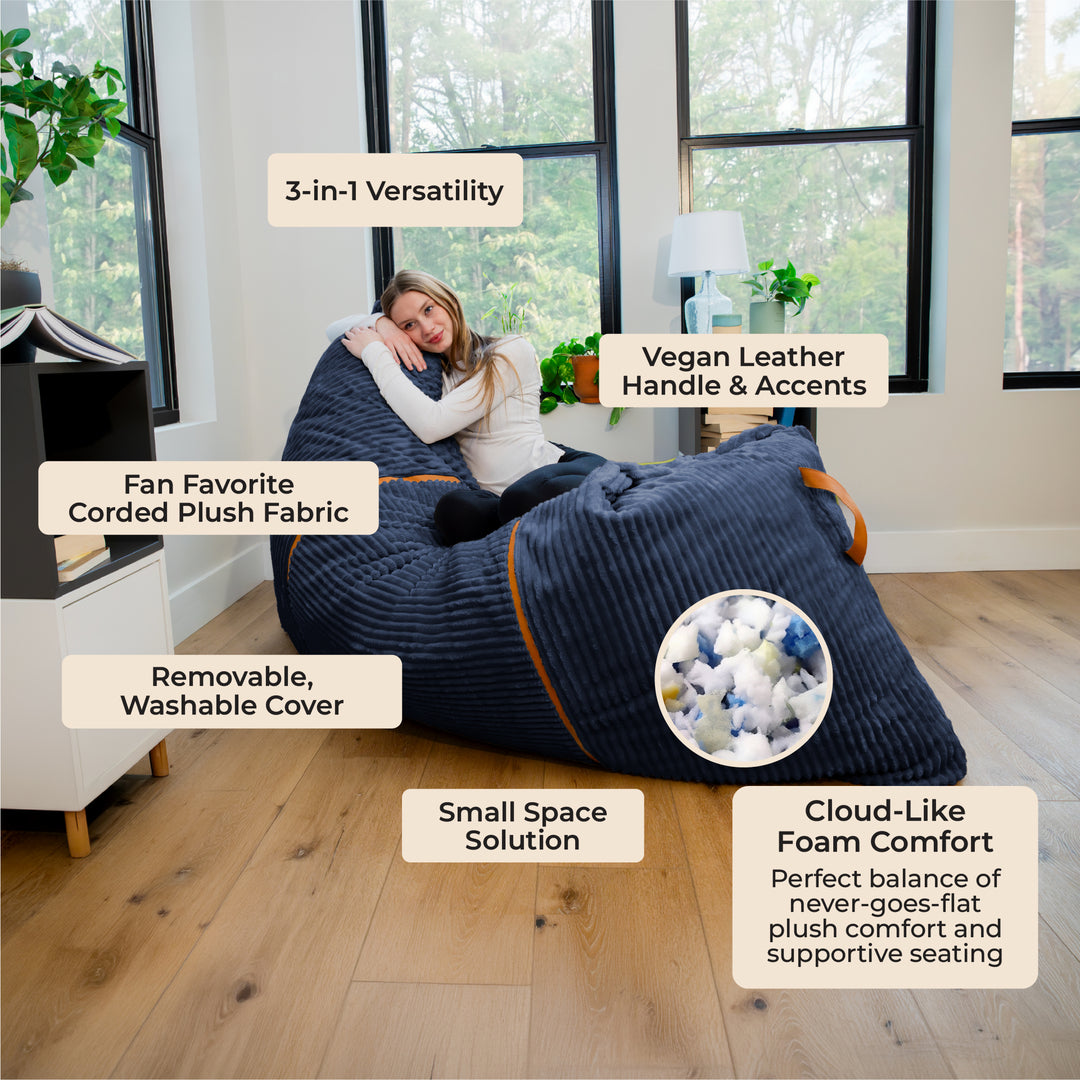 A woman reclines on a large ribbed bean bag chair in a bright living room. The bean bag is dark navy blue with orange trim and a circular inset showing the plush foam interior. 