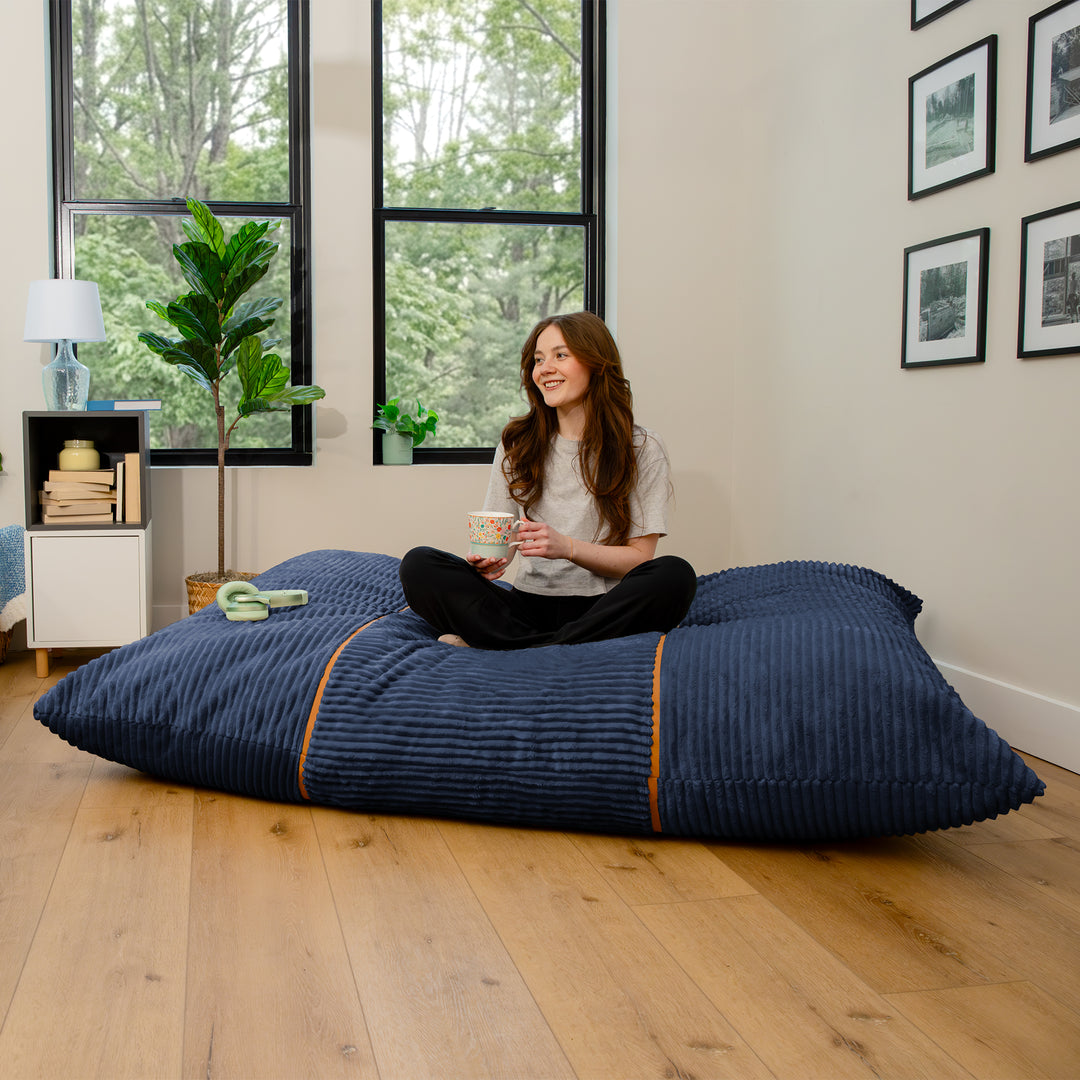 A woman sits cross-legged on a large navy blue ribbed floor cushion in a bright room with wood floors, large windows, and framed wall art. 