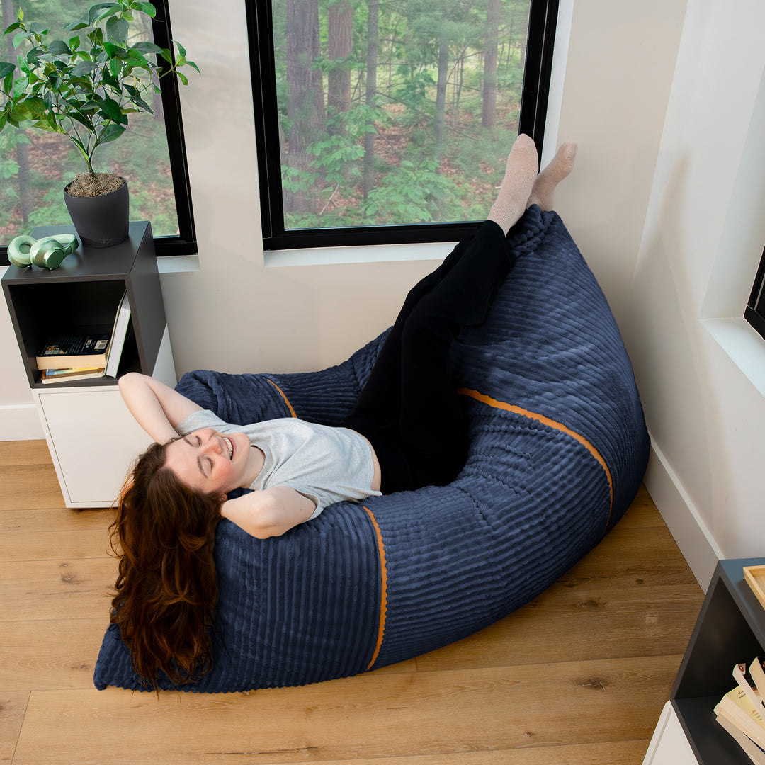 A woman relaxes on a large navy blue bean bag chair with orange piping, set in a modern room with wood floors and large windows. 