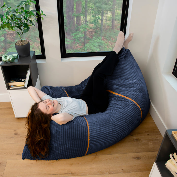 A woman relaxes on a large navy blue bean bag chair with orange piping, set in a modern room with wood floors and large windows. 