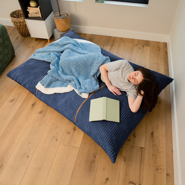 A person lies on a large dark blue floor cushion or mattress in a bedroom, covered with a light blue blanket, with a book in front of them. The room has light wood flooring and neutral walls. 