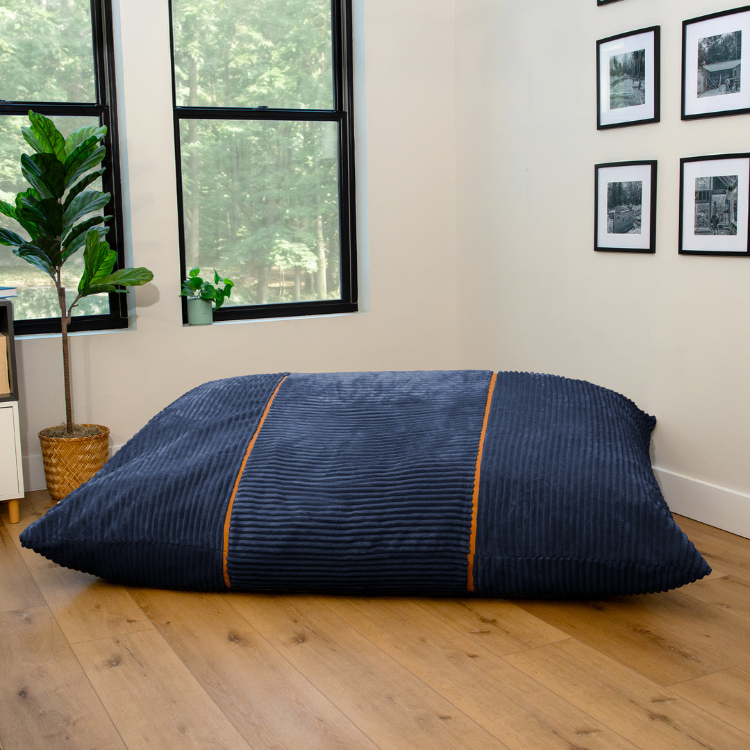 A large rectangular floor cushion or lounger in dark navy blue corduroy fabric with two vertical orange stripes, placed on a light wood floor in a modern room with windows and framed wall art. 