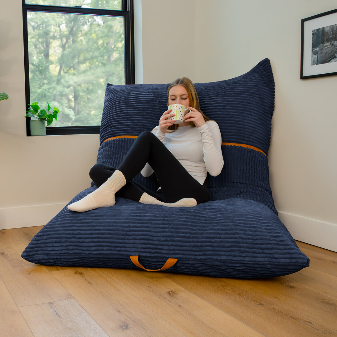 A woman sits cross-legged on a large navy blue ribbed floor cushion with matching back pillows in a bright room with wood floors and a window. 