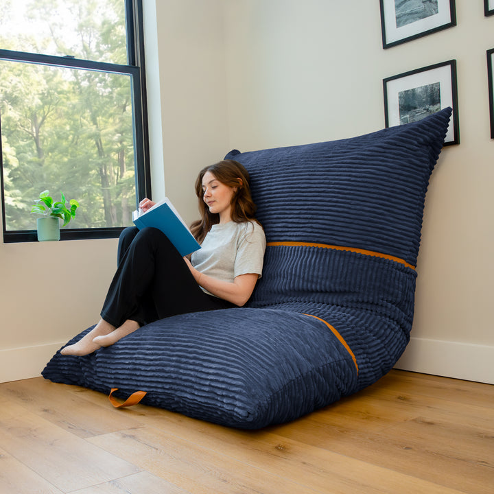 A woman sits on a large navy blue ribbed bean bag chair in a bright room with a window, wooden floor, and framed wall art. 