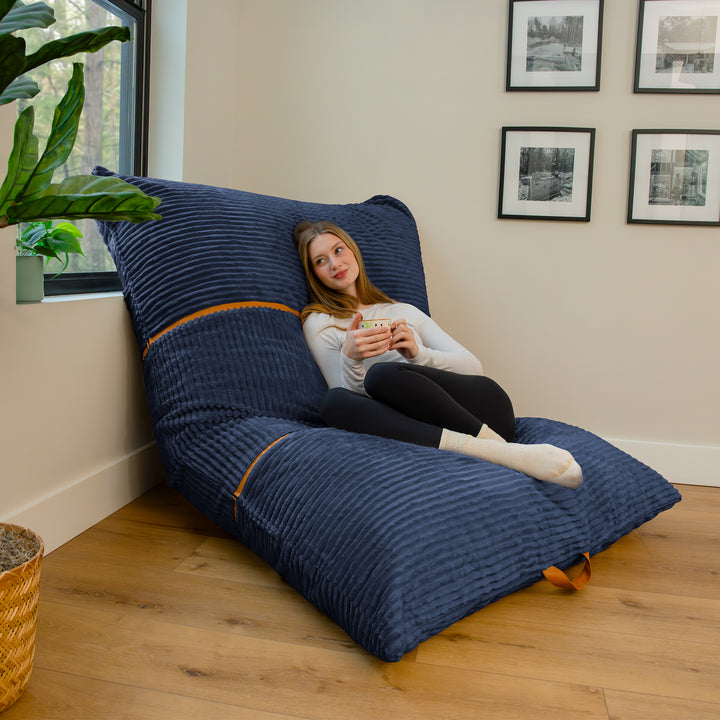 A woman reclines on a large navy blue ribbed floor lounger in a bright room with wood floors, a window, framed wall art, and a potted plant. 