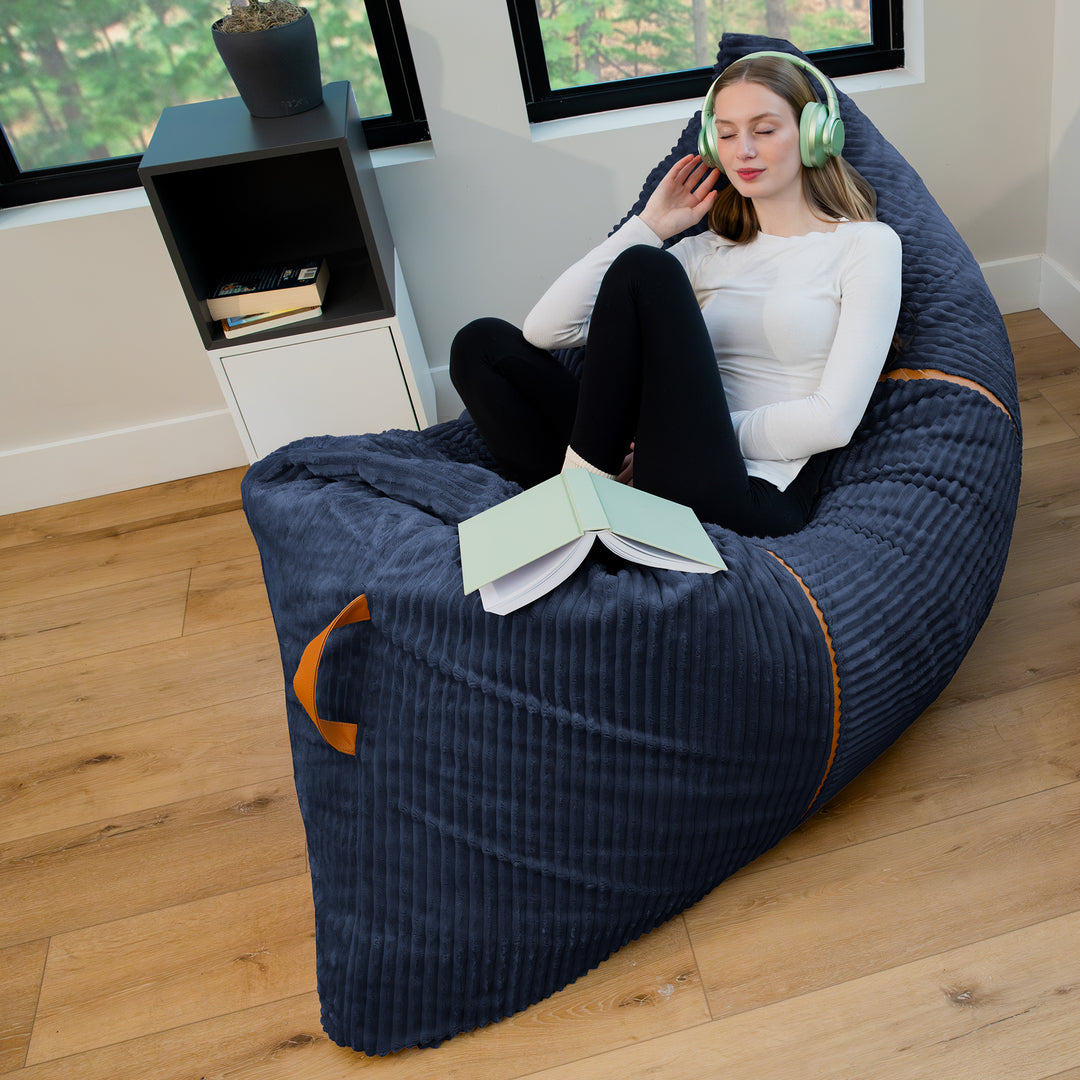 A woman relaxes in a large navy blue ribbed bean bag chair with orange accents, sitting in a bright room with wood floors and windows. 