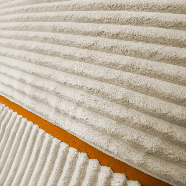 Close-up of a ribbed corduroy fabric in warm off-white, with a diagonal orange accent stripe visible along the lower left. 