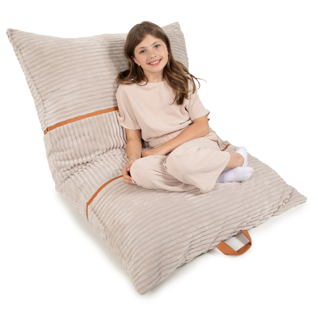A young girl with long brown hair sits cross-legged on a large, adjustable floor cushion. The cushion is made of a ribbed beige fabric with a brown leather strap handle and a matching brown stripe detail. 