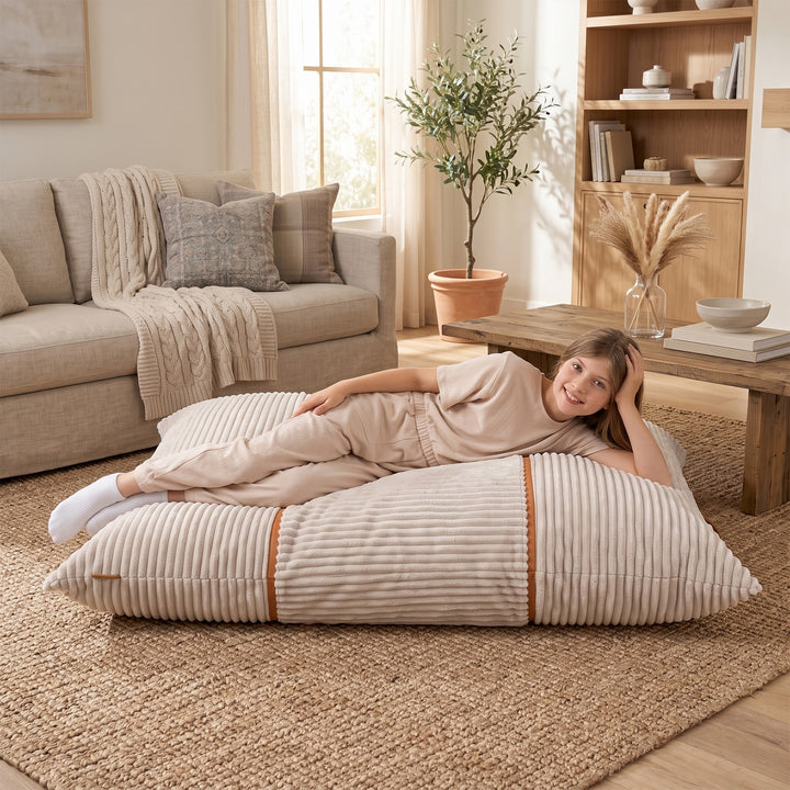 A woman in beige loungewear reclines on a large ribbed floor pillow in a bright, modern living room with a sofa, coffee table, and bookshelf. 