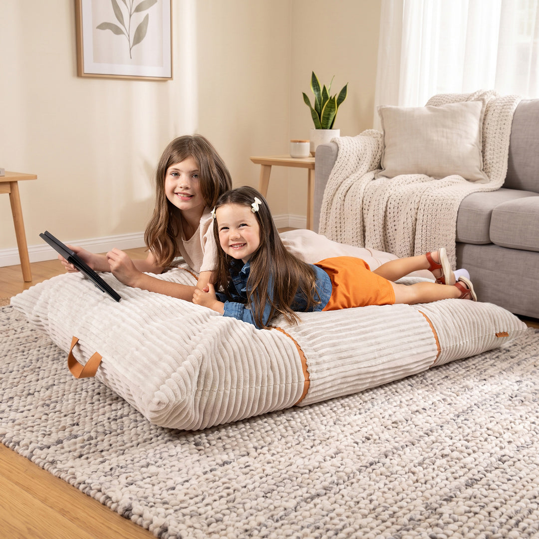 Two young girls lying on a large cream ribbed floor cushion in a cozy living room setting. The cushion is placed on a textured gray rug in front of a gray sofa with a knit throw blanket and a potted plant on a side table. 