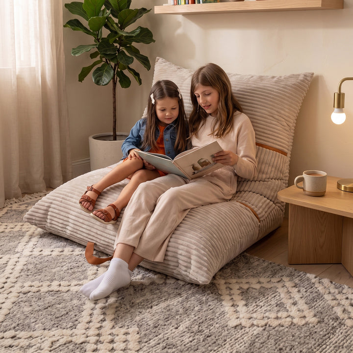 A woman and a child relax on a large striped beige floor cushion in a cozy room, reading a book together. The scene includes a wooden side table with a mug, a potted plant, and warm ambient lighting. 