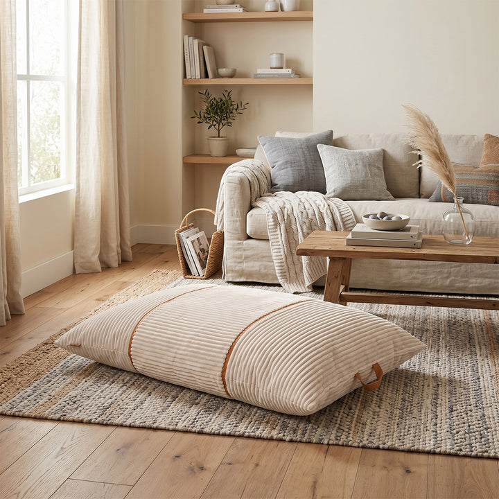 A large rectangular floor cushion with a beige and white striped corduroy-like texture and brown trim, styled in a bright, modern living room on a woven rug. 
