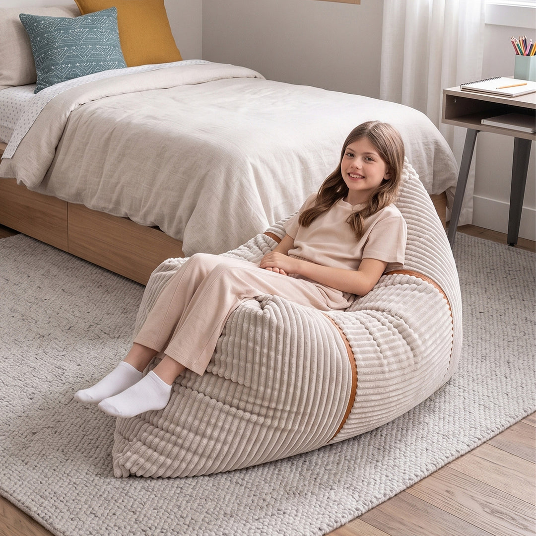A young girl wearing a beige short-sleeve top and matching pants sits on a large ribbed corduroy bean bag chair in a cozy bedroom setting. 
